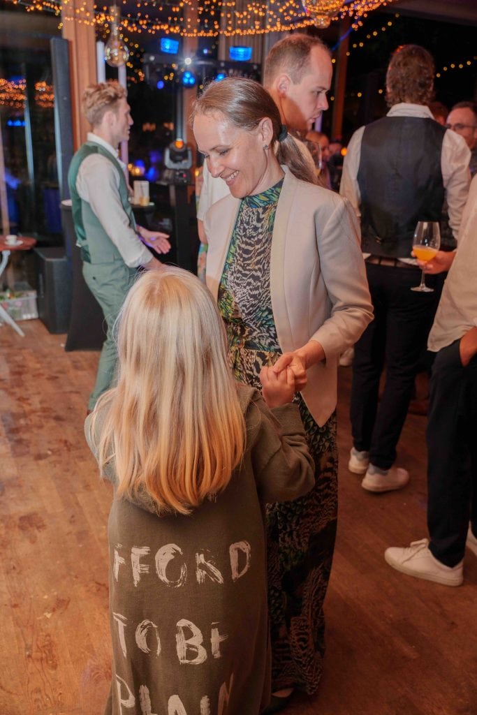 Frau tanzt mit Kind bei festlicher Veranstaltung, umgeben von Gästen und Lichterdekorationen.