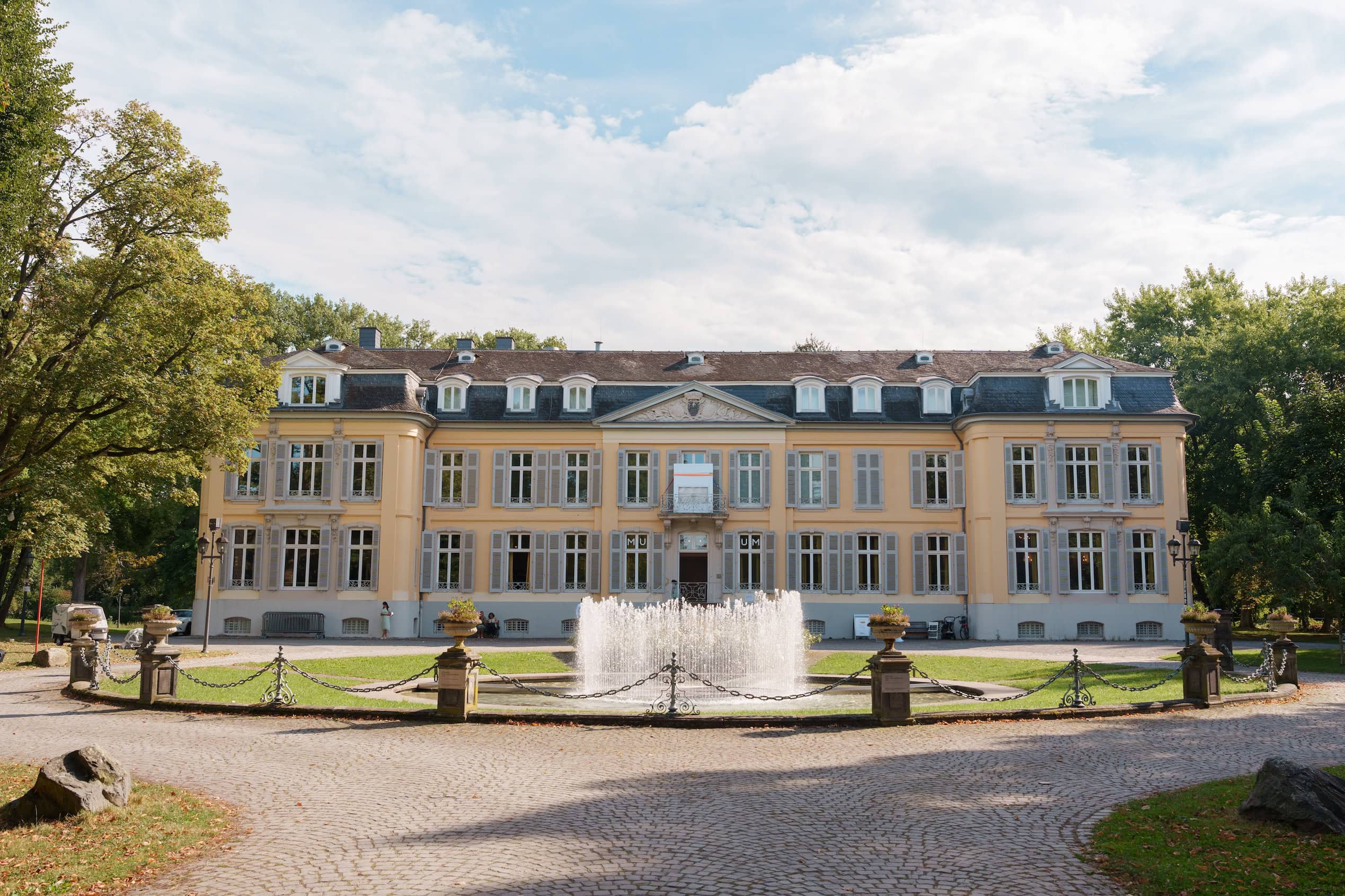 Historisches Schloss mit Springbrunnen im Park, umgeben von Bäumen, bei schönem Wetter.